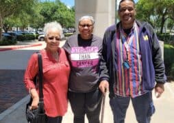 Three adults stand smiling outdoors on a sunny day. The person in the center, living with metastatic breast cancer, wears a headscarf and a "Stronger Than Cancer" shirt as their companions stand close, showing support. Trees and parked cars are behind them.