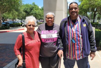 Three adults stand smiling outdoors on a sunny day. The person in the center, living with metastatic breast cancer, wears a headscarf and a "Stronger Than Cancer" shirt as their companions stand close, showing support. Trees and parked cars are behind them.