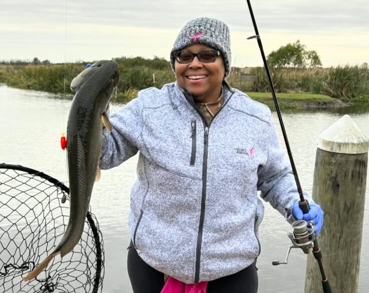 A smiling person in a gray jacket and beanie holds a fishing rod in one hand and a large fish in the other, standing by the water with reeds and cloudy sky in the background.