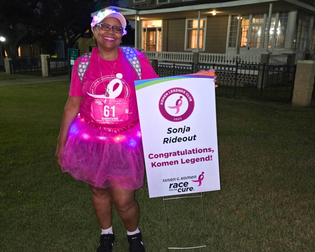 A smiling woman in a pink t-shirt, tutu, and glowing accessories stands outside at night holding a sign that celebrates her as a "Komen Legend" and breast cancer survivor at a Race for the Cure event.