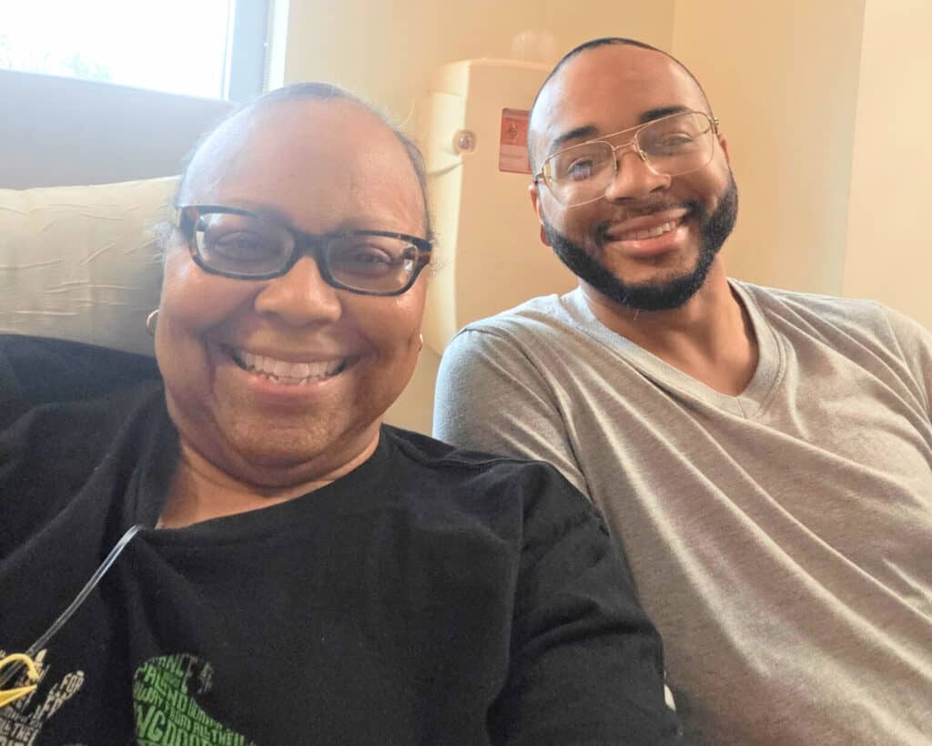 Two people are smiling while sitting indoors. The person on the left is wearing glasses and a black shirt, and the person on the right has a beard, glasses, and a gray shirt. They appear happy and relaxed together.