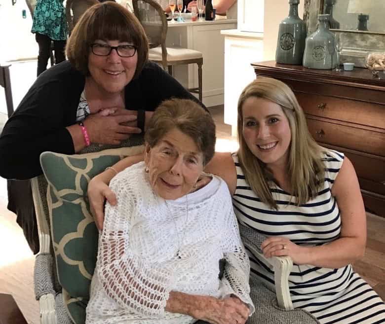 Three women pose together indoors. An elderly woman in a white sweater sits between a younger woman in a striped dress and another woman leaning over the chair, all smiling warmly at the camera.