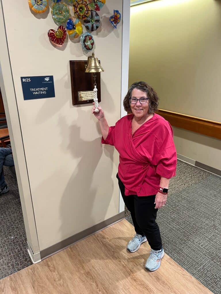 A breast cancer survivor in a pink top smiles while ringing a bell in a medical facility next to a sign that reads “Treatment Waiting.” Colorful glass art decorates the wall above the bell, celebrating her completion of radiation.