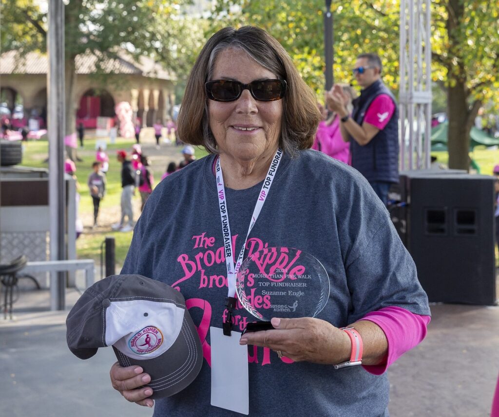 Smiling woman wearing sunglasses and a race shirt holds a trophy and a baseball cap at an outdoor breast cancer fundraiser, with people and trees in the background.
