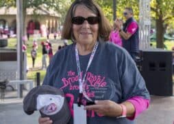 Smiling woman wearing sunglasses and a race shirt holds a trophy and a baseball cap at an outdoor breast cancer fundraiser, with people and trees in the background.