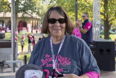 Smiling woman wearing sunglasses and a race shirt holds a trophy and a baseball cap at an outdoor breast cancer fundraiser, with people and trees in the background.