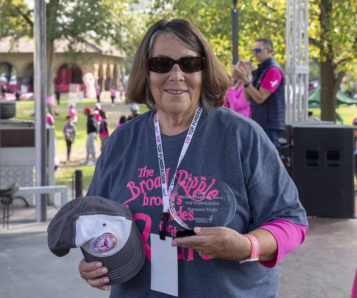 Smiling woman wearing sunglasses and a race shirt holds a trophy and a baseball cap at an outdoor breast cancer fundraiser, with people and trees in the background.