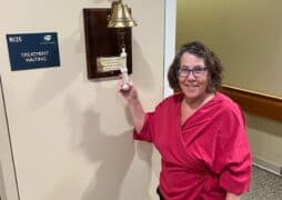 A breast cancer survivor in a pink top smiles while ringing a bell in a medical facility next to a sign that reads “Treatment Waiting.”