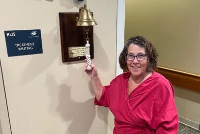 A breast cancer survivor in a pink top smiles while ringing a bell in a medical facility next to a sign that reads “Treatment Waiting.”