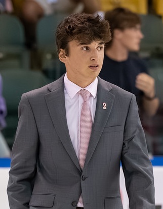 A young man in a gray suit and pink tie stands indoors, looking to the side. He has brown hair and wears a pink breast cancer awareness ribbon pin on his jacket. Blurred people and seats are visible in the background.