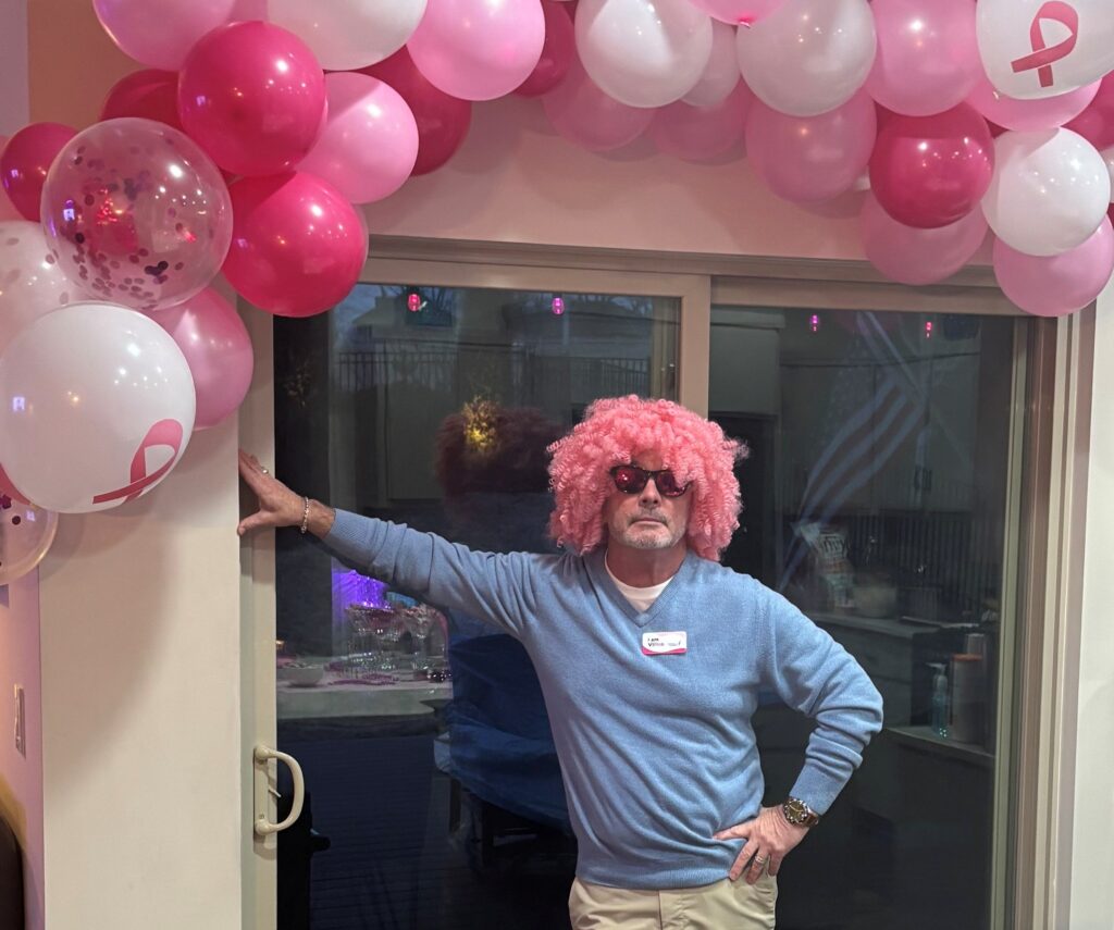 A man poses under a pink balloon arch. He is wearing a blue shirt, sunglasses and pink curly wig as a part of his participation in Komen's VIPink ambassador fundraising campaign.