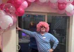 A man poses under a pink balloon arch. He is wearing a blue shirt, sunglasses and pink curly wig as a part of his participation in Komen's VIPink ambassador fundraising campaign.