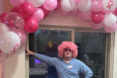A man poses under a pink balloon arch. He is wearing a blue shirt, sunglasses and pink curly wig as a part of his participation in Komen's VIPink ambassador fundraising campaign.