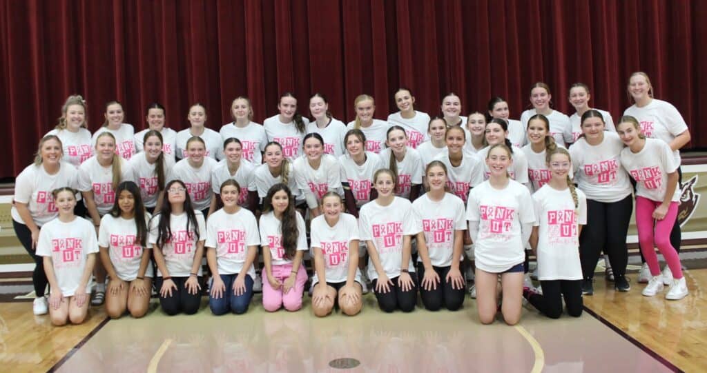 A large group of girls poses indoors in front of red curtains, all wearing white "Pink Out" T-shirts for a volleyball breast cancer fundraiser, smiling at the camera. They are arranged in two rows on a gym floor.