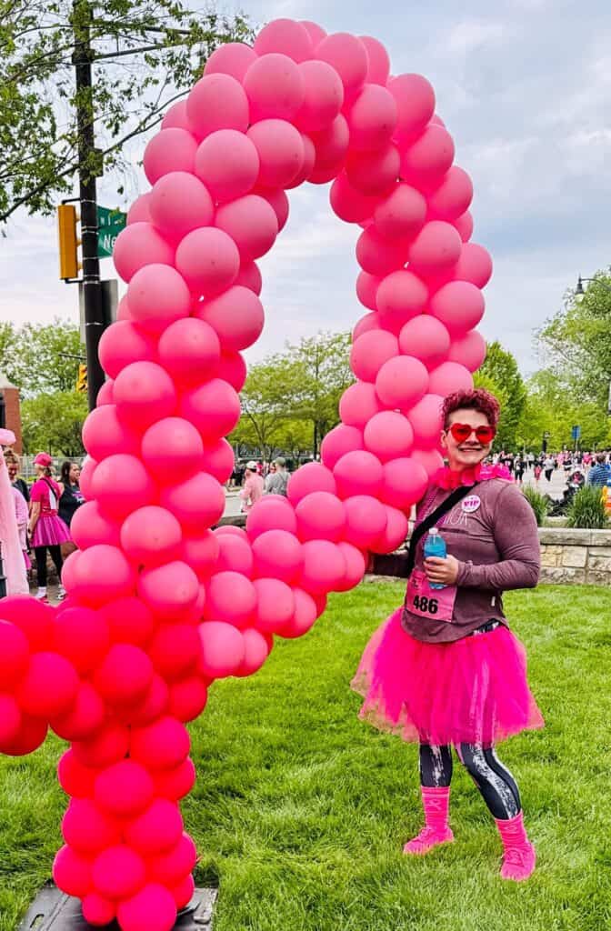 A woman in a pink tutu stands by a large pink ribbon balloon sculpture at a breast cancer fundraiser event.