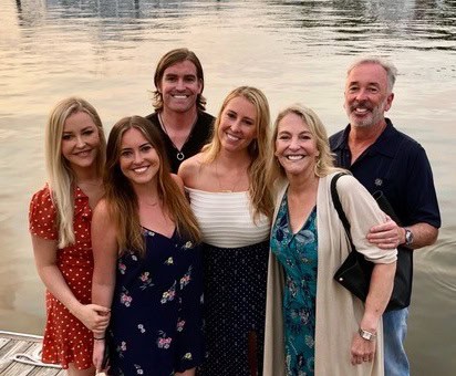 Six adults smile together by a waterfront, standing close on a dock with calm water in the background. The group includes four women in dresses and two men, all appearing happy and relaxed outdoors, showing the importance of connection and support for those affected by metastatic breast cancer.