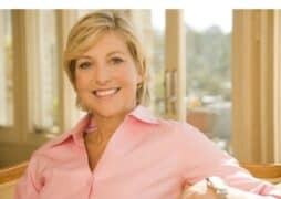 A woman with short blonde hair, wearing a light pink shirt, smiles while sitting indoors near large windows with sunlight streaming in.