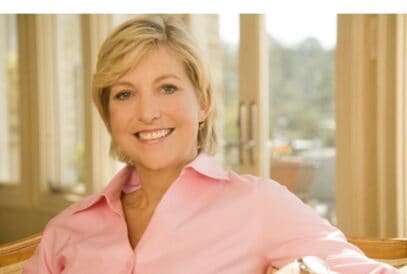 A woman with short blonde hair, wearing a light pink shirt, smiles while sitting indoors near large windows with sunlight streaming in.