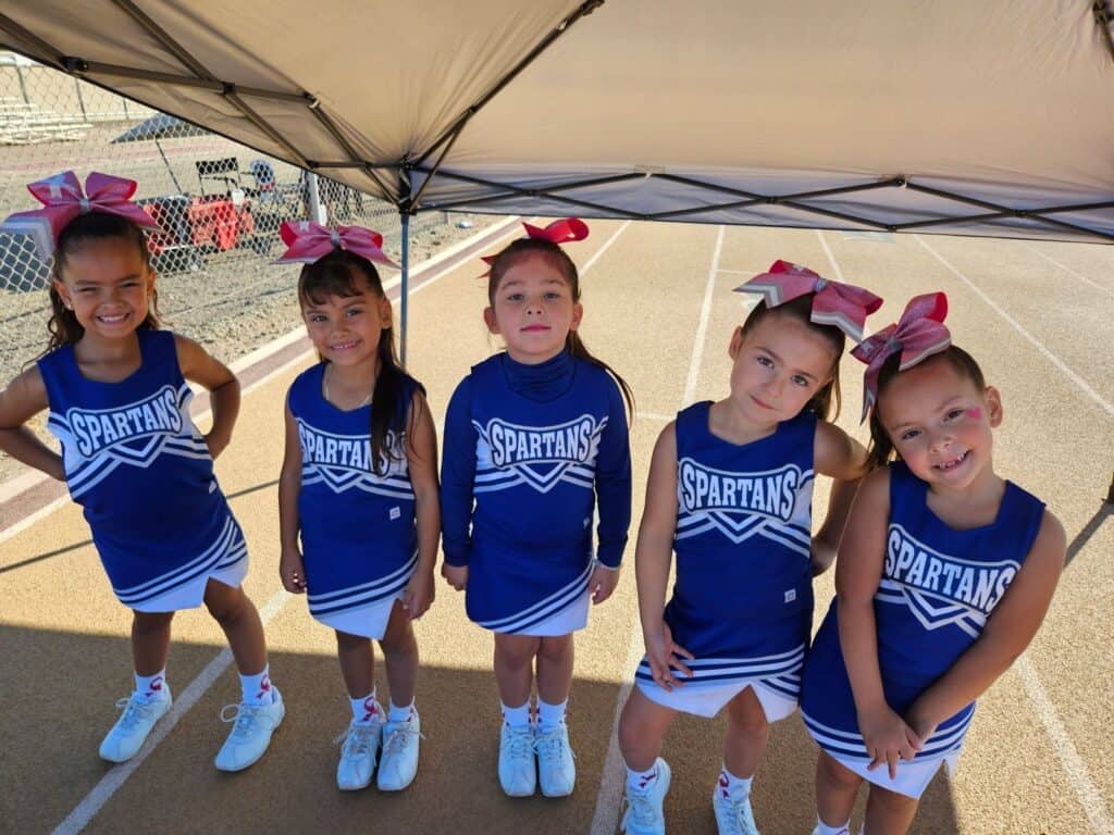 Five young girls in matching blue "Spartans" cheerleading uniforms and pink bows stand in a row under a canopy on a track, smiling and posing for the camera during a youth sports breast cancer fundraiser.