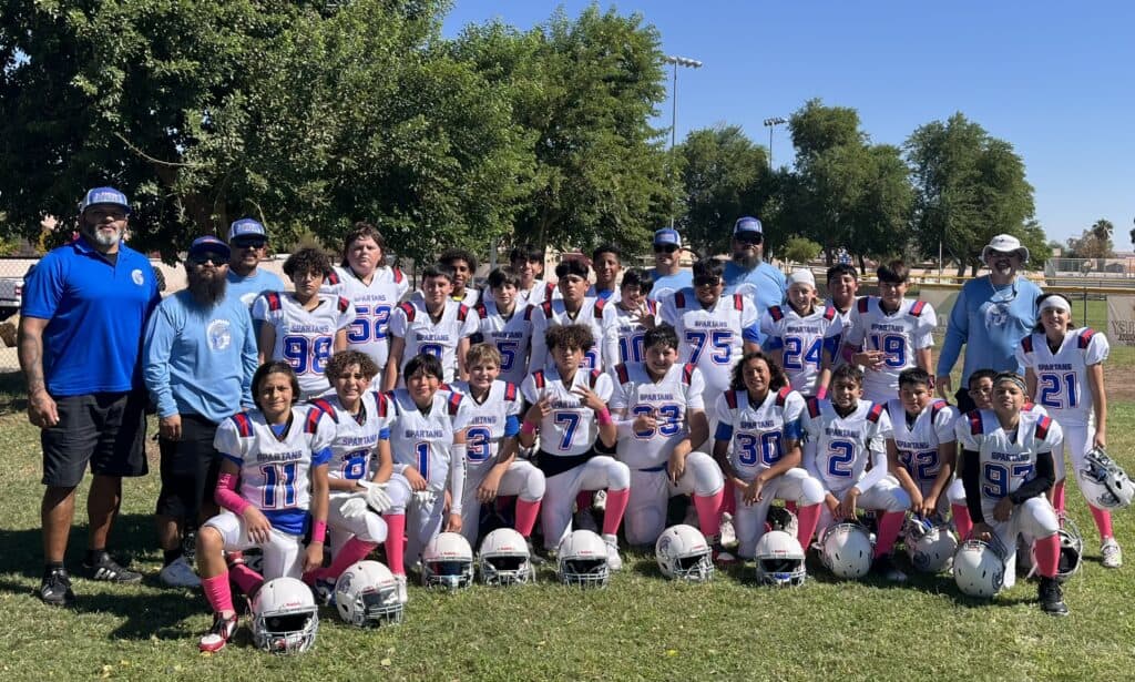 A youth football team in white, pink and blue uniforms poses for a group photo outdoors with their coaches at a youth sports breast cancer fundraiser. Helmets are on the grass, with trees and a clear blue sky in the background.