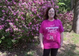 A breast cancer survivor with medium-length brown hair stands outdoors in front of flowering pink bushes, smiling. She wears glasses and a bright pink “Stronger Together” Race for the Cure event T-shirt with a race bib numbered 35.