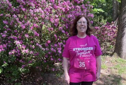 A breast cancer survivor with medium-length brown hair stands outdoors in front of flowering pink bushes, smiling. She wears glasses and a bright pink “Stronger Together” Race for the Cure event T-shirt with a race bib numbered 35.