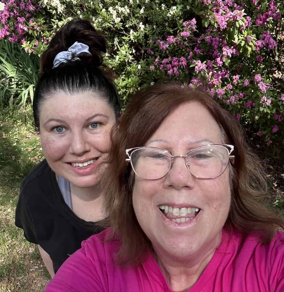 A woman with medium length brown hair and glasses smiles at the camera. Another woman stands behind her, smiling, her hair pulled into a ponytail.